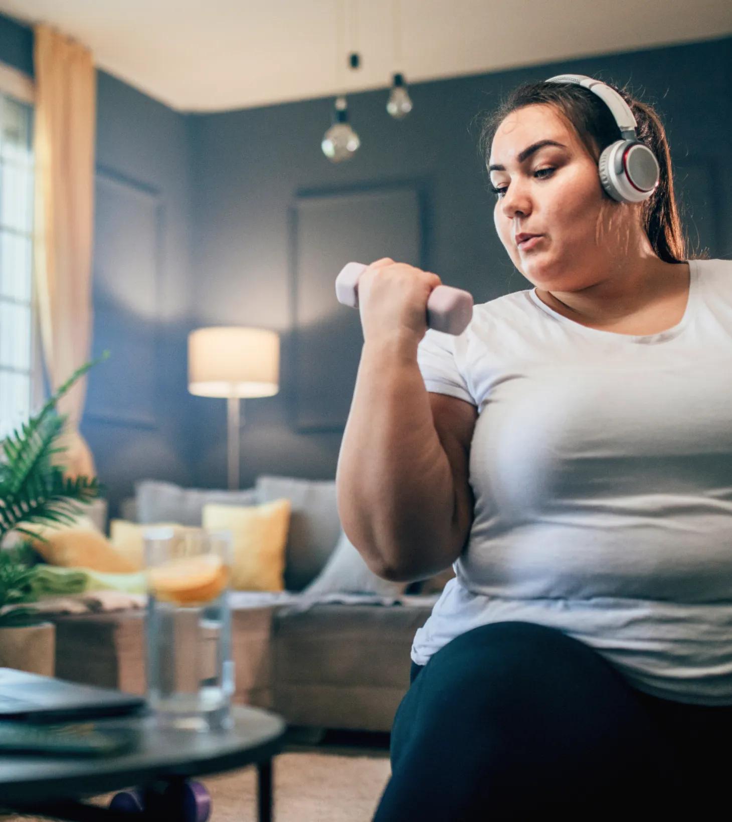 An obese woman in her early thirties doing bicep curls at home with water nearby. She’s sticking to the convenient activity plan she created with her virtual program care team.