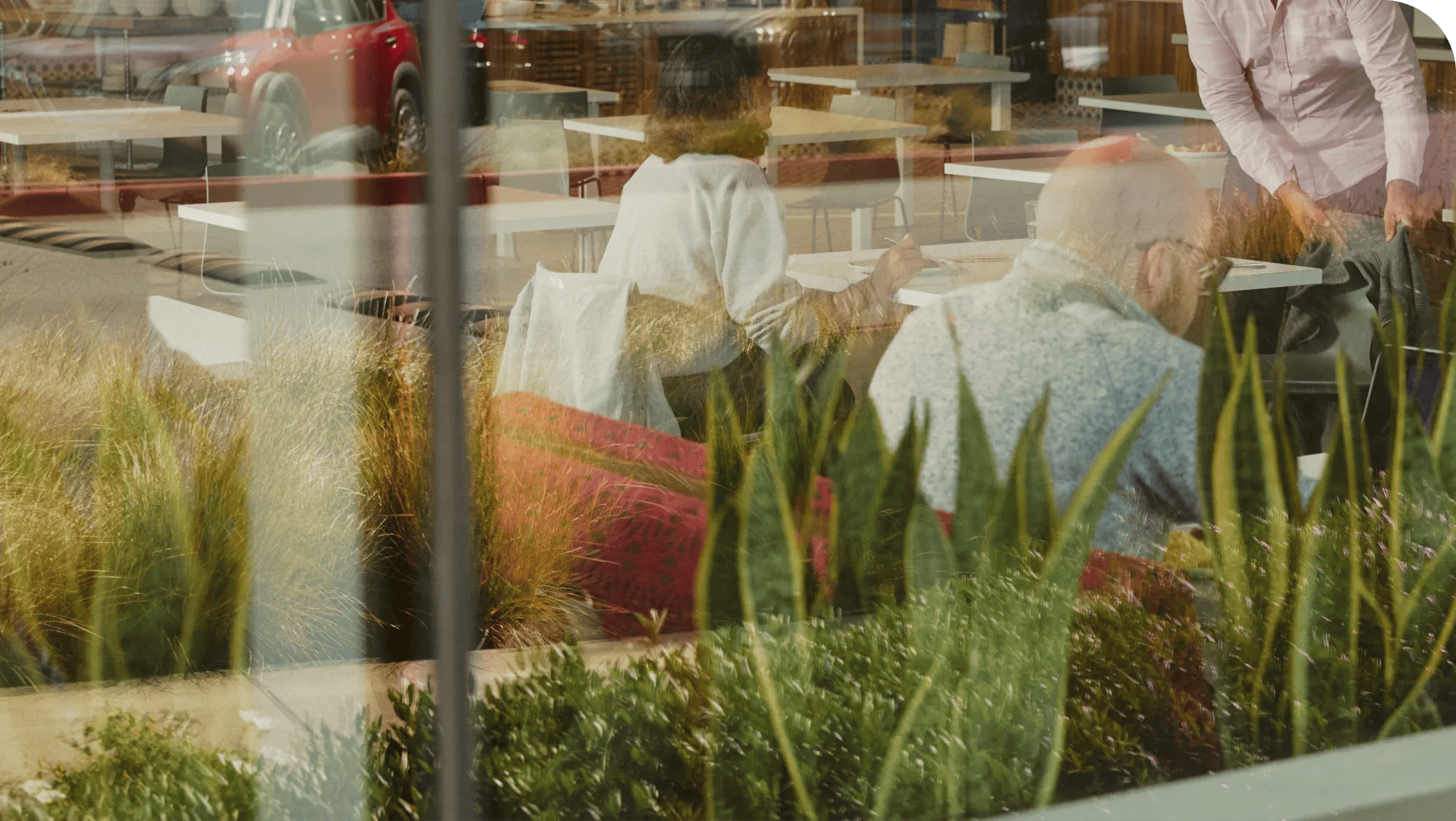 People sitting at tables inside a Verily Cafetaria, viewed through a glass window framed by green plants. 