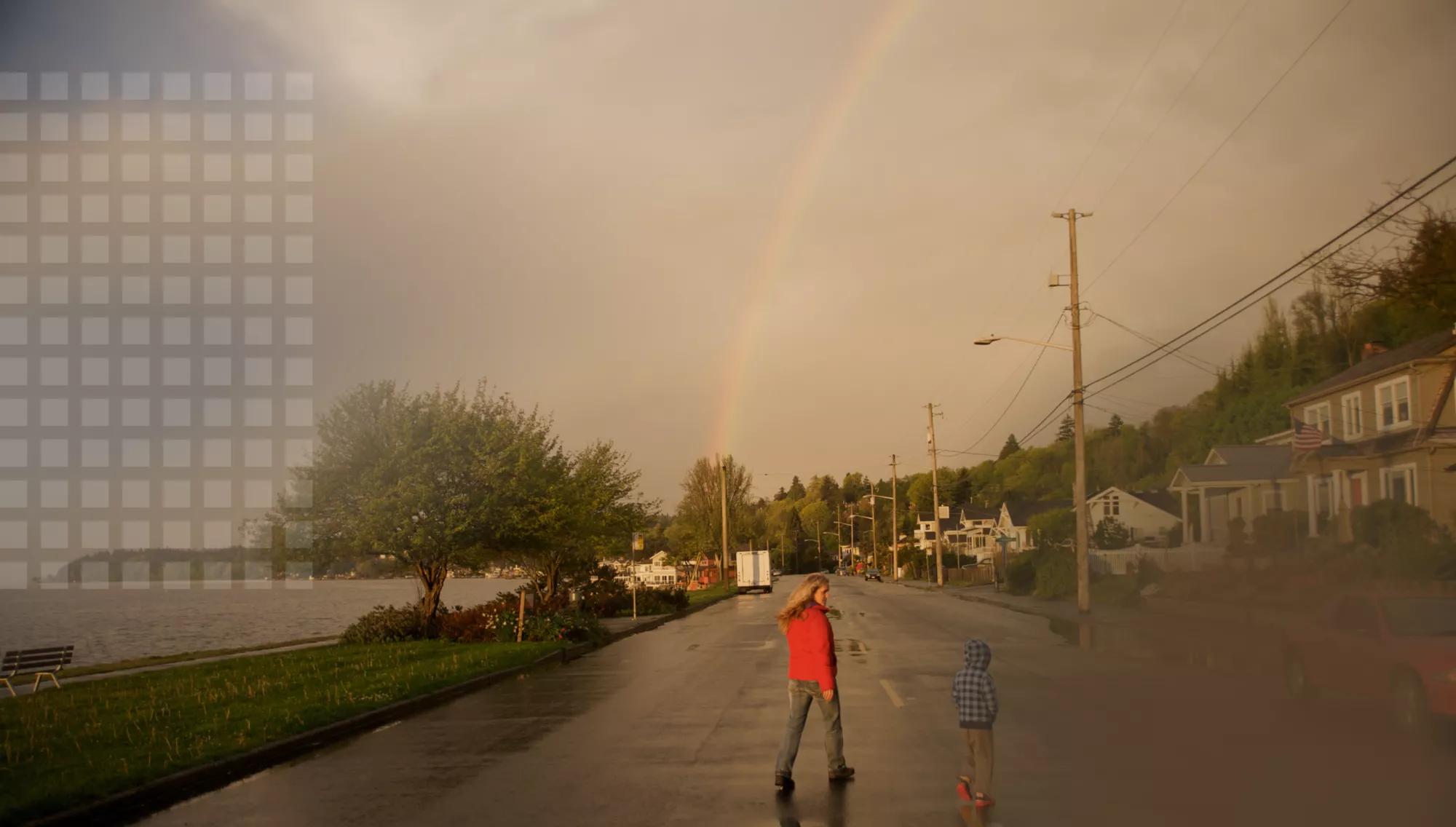 Mother and son walking with zest in their community along the water on a drizzly day with a rainbow on the horizon.